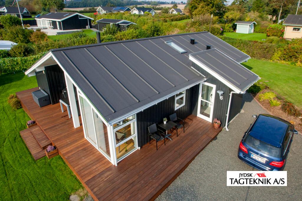 Oppefrar vist parcelhus i træ med mørkt tag, træterrasse og en bil parkeret i indkørslen.